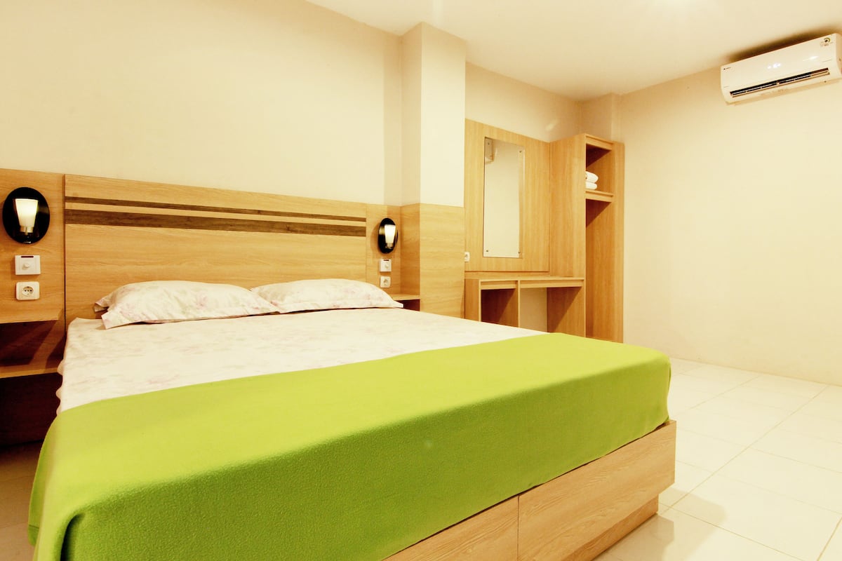 Una cama bien hecha con una colcha verde, cabecero de madera y lámparas de noche.