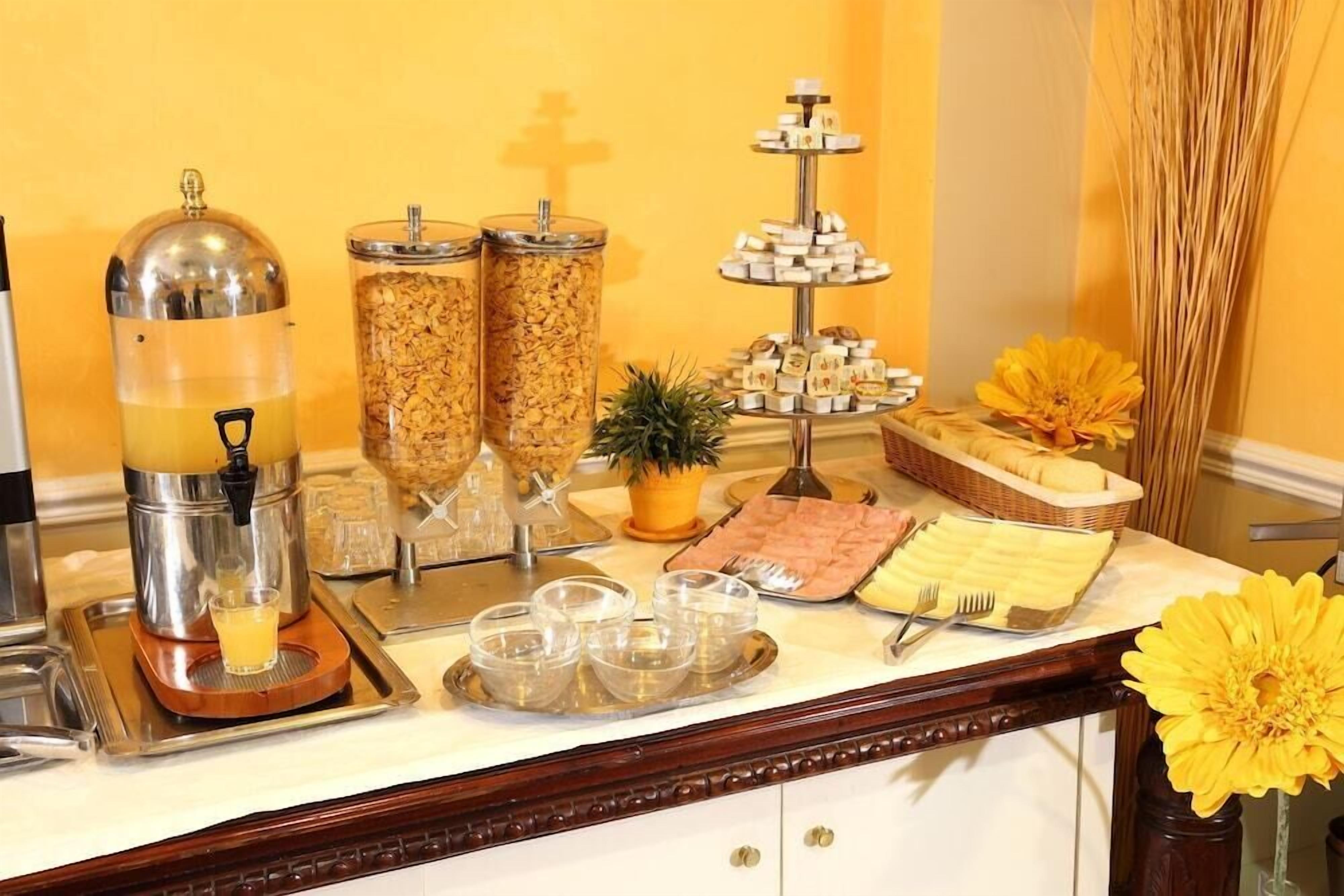 Montaje de desayuno buffet con dispensador de jugo, recipientes de cereal, un expositor de postres escalonado y un arreglo floral.