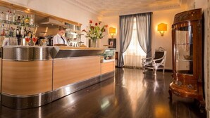 Un espace bar avec un comptoir en bois poli, un barman et une variété de bouteilles. On y trouve un vase décoratif avec des fleurs, un fauteuil classique et un meuble ancien.