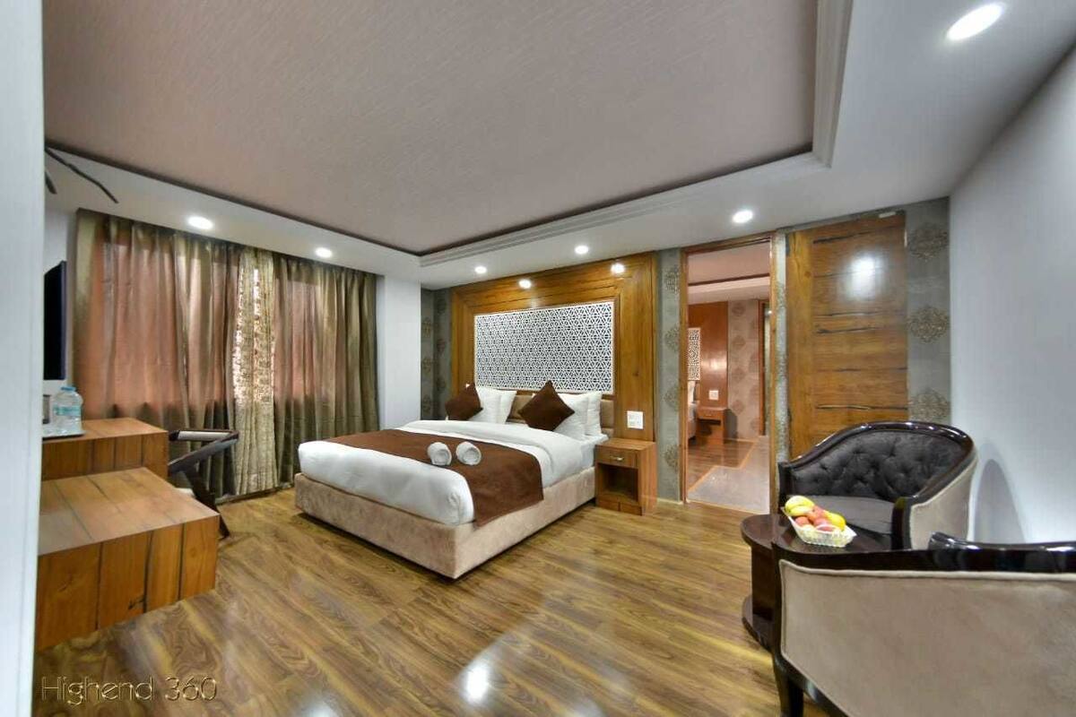 Quarto de hotel moderno com chão de madeira, uma cama com cabeceira, mesa de cabeceira, uma cadeira e uma pequena mesa com frutas.