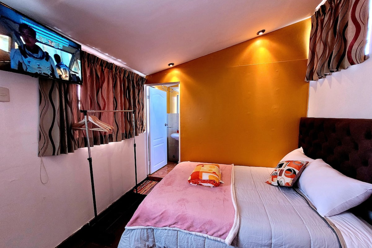A bedroom with a bed, a TV mounted on the wall, a bathroom visible through an open door, and a patterned curtain.