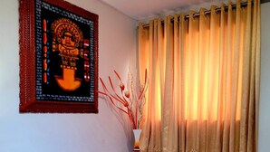Una habitación con una mesa redonda de cristal, dos rincones de estar con cojines, un cuadro colgado en la pared, una cortina y un jarrón con ramas decorativas.