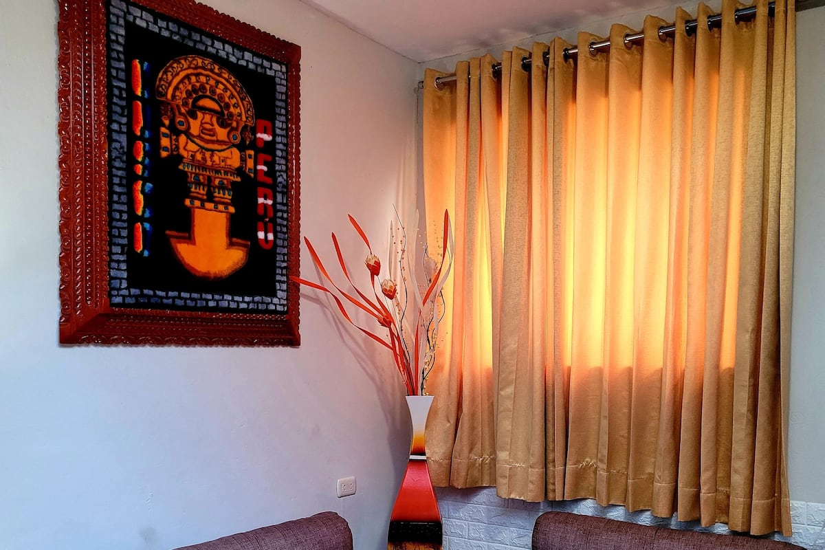 A room with a round glass table, two cushioned seating areas, a wall-mounted painting, a curtain, and a vase with decorative branches.