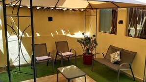 Un espacio exterior techado con sillas, una mesa, una planta en maceta y una ventana con cortinas.