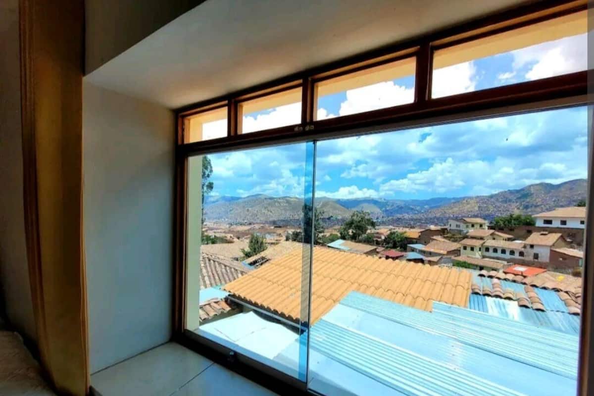 A room with a large window offering a view of a town with terracotta-roofed buildings and distant mountains.