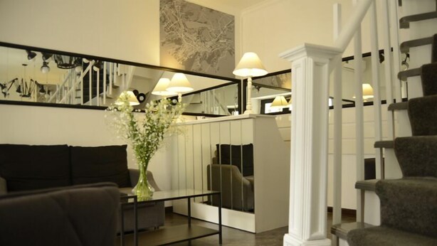 A modern living room with a sofa, a coffee table, and a vase of flowers. There is a staircase with a railing and a wall-mounted mirror. - Hotel Opera (Tbilisi)