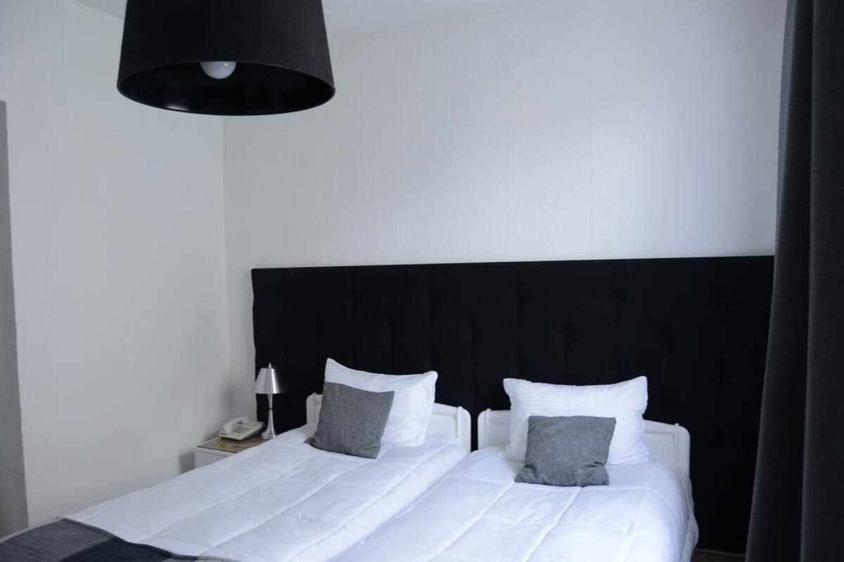 A hotel room with a black headboard, two beds with white bedding and gray pillows, a bedside table with a lamp, and a phone.