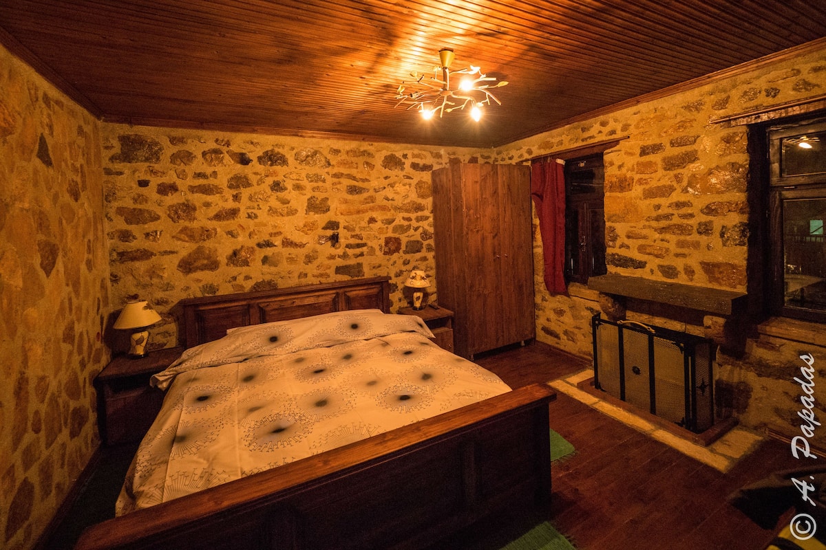 A room with a stone wall, wooden ceiling, a bed with a patterned bedspread, a nightstand with a lamp, and a small table with a chair.