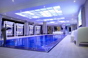 Un área de piscina interior moderna con techo transparente, sillas reclinables blancas y piso de baldosas.