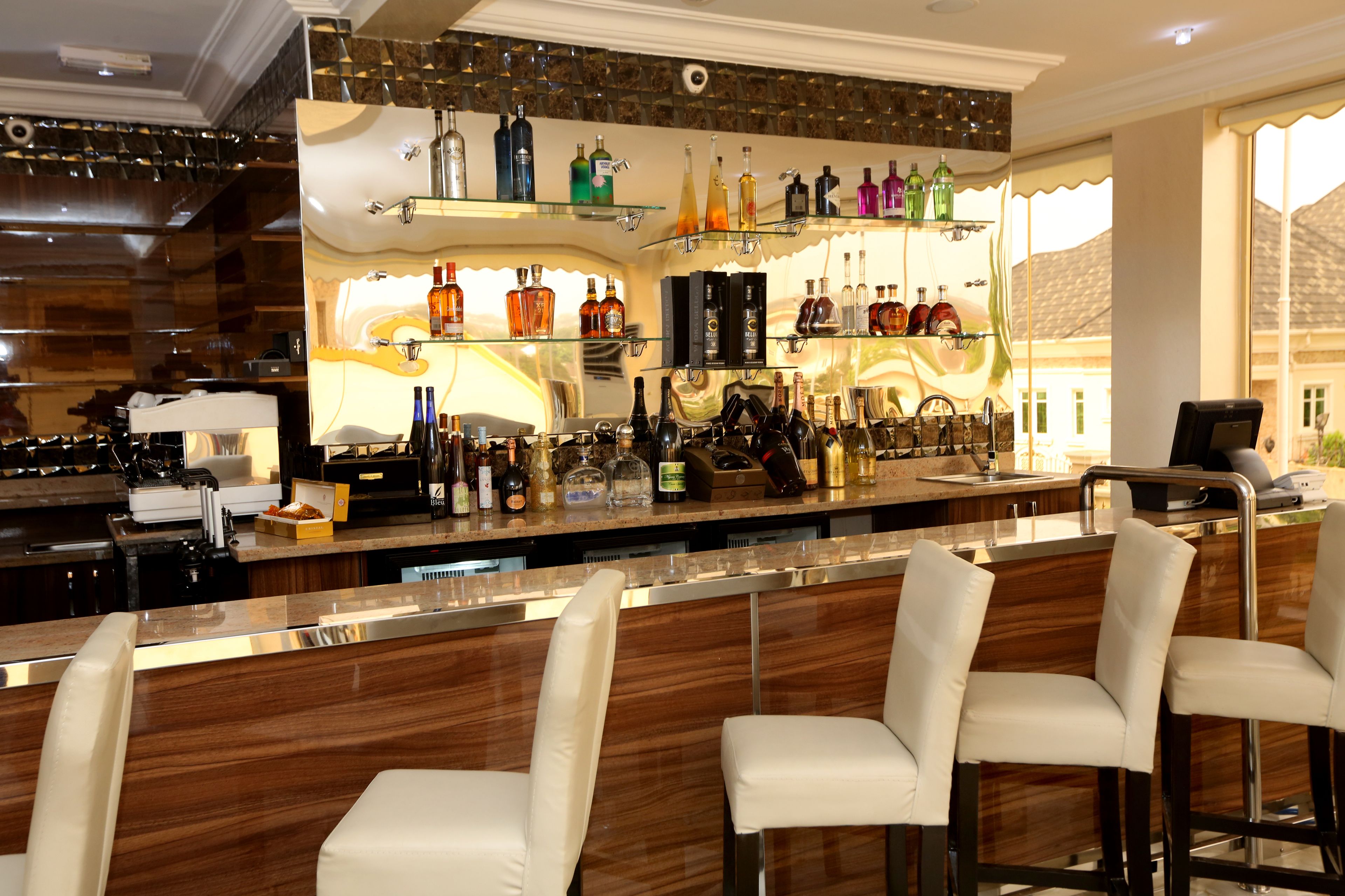 Een bar met een houten toonbank, diverse drankflessen en witte, beklede barstoelen.