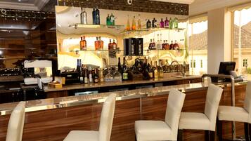 Een bar met een houten toonbank, diverse drankflessen en witte, beklede barstoelen.