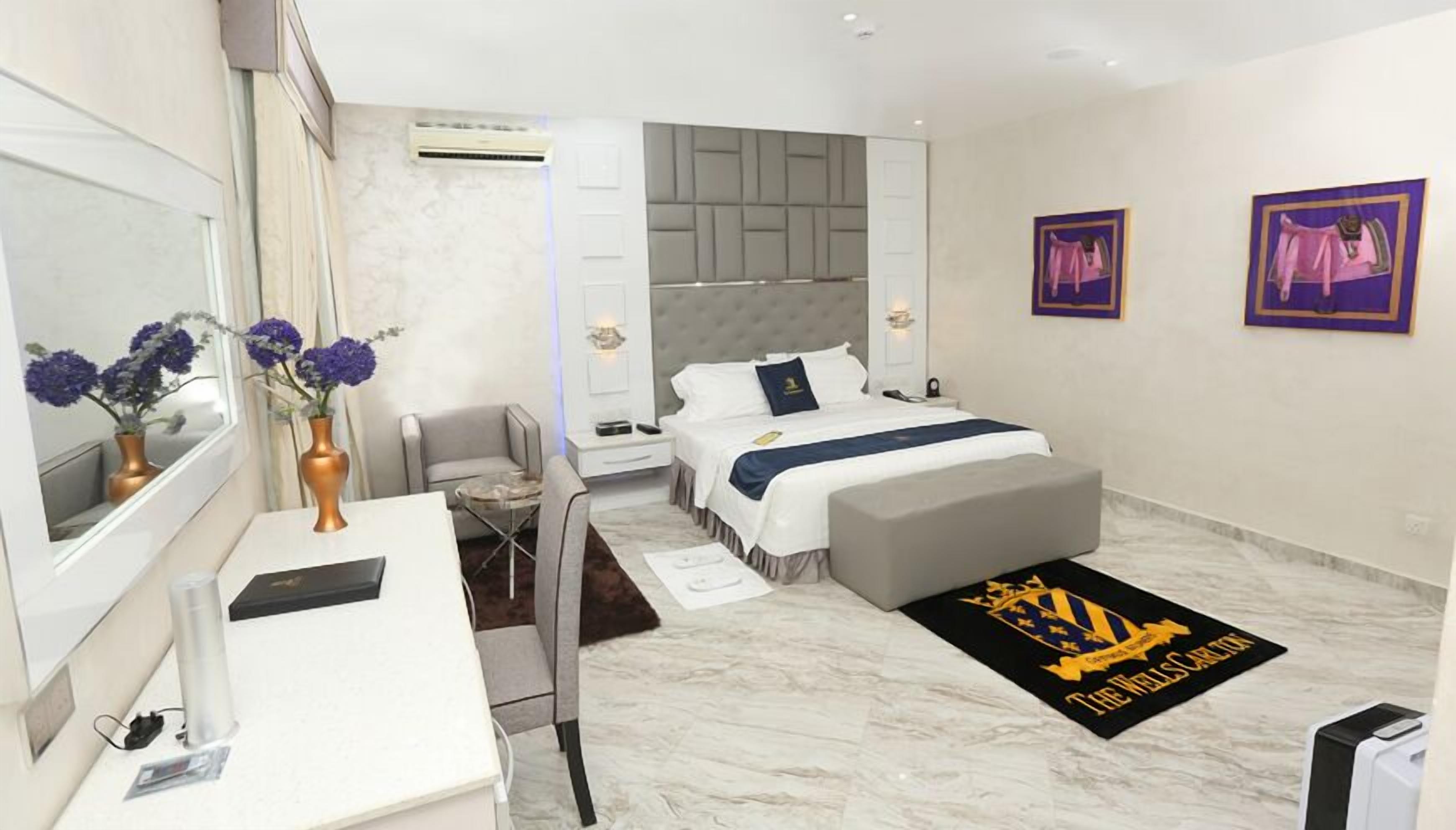 A modern hotel room with a bed, a desk, a chair, a small table, and a sofa. There are two paintings on the wall and a rug on the floor.