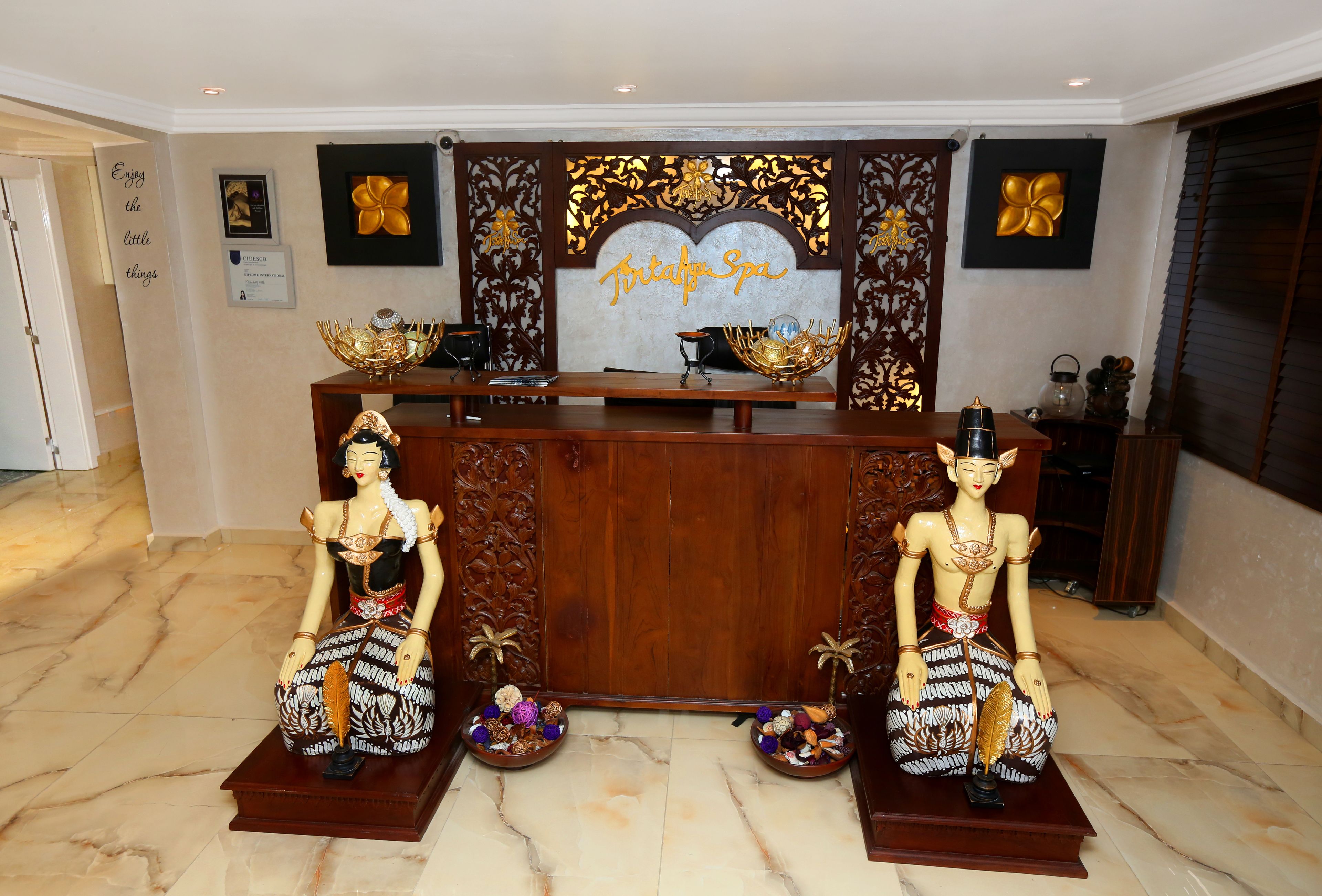 Un comptoir d’accueil orné de deux statues décoratives, une enseigne indiquant « Trinity Spa » et un tableau encadré représentant une fleur.