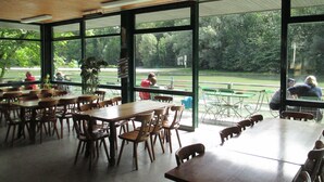 An indoor dining area with wooden tables and chairs, large windows offering views of a park and a road. - Munich Camping Glamping and Dorms (Munich)