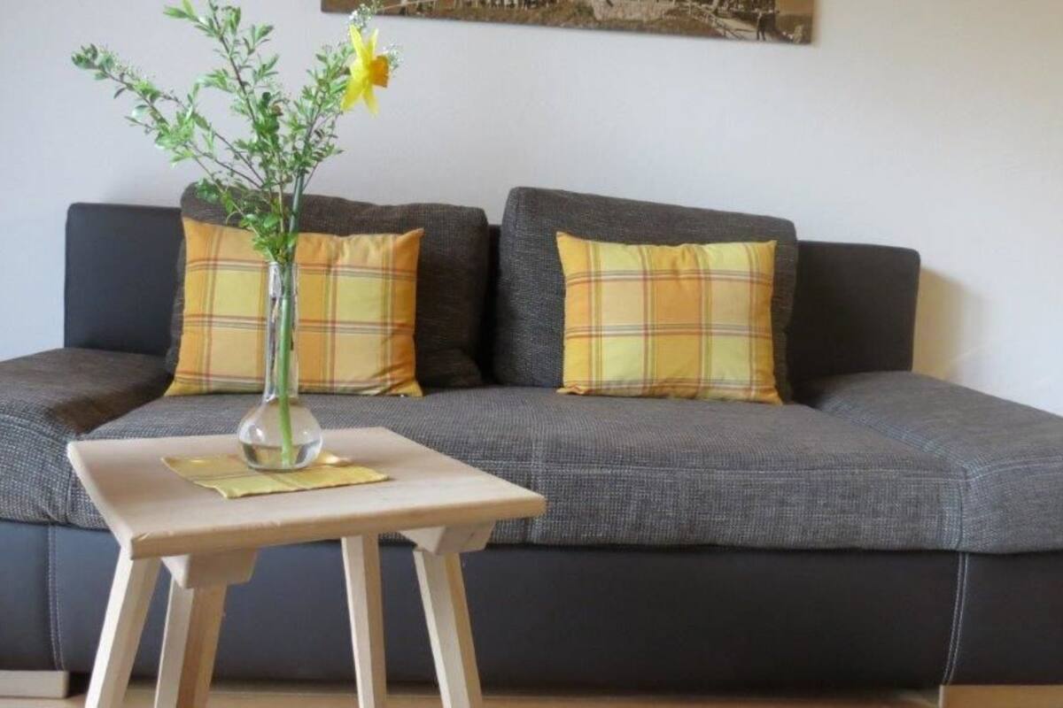 Um sofá cinzento com almofadas xadrez, uma pequena mesa de madeira com um vaso de flores e um quadro na parede.