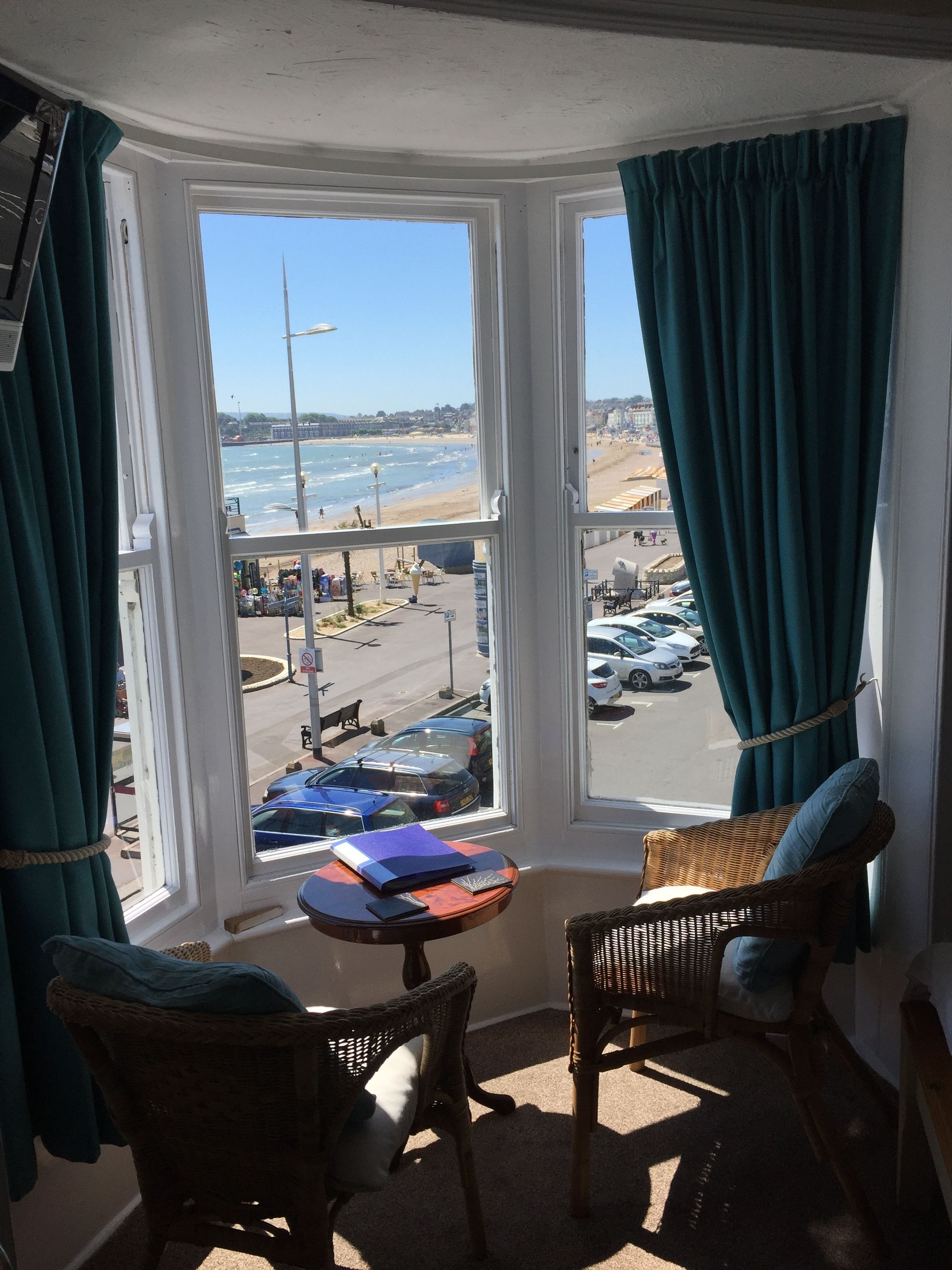 Ein Zimmer mit Erkerfenster, das Blick auf Strand und Parkplatz bietet. Korbstühle und ein kleiner Tisch mit Büchern und einer Lampe stehen bereit.