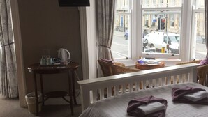Une chambre d’hôtel avec un lit, un téléviseur fixé au mur, une fenêtre avec des rideaux, une petite table avec une bouilloire et une vue sur la rue.