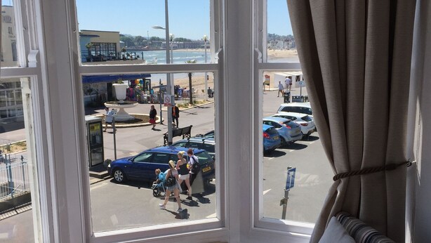 Une pièce dotée d’une grande baie vitrée donnant sur une rue animée et une plage. On y trouve un fauteuil à rayures, une petite table ronde, et une vue sur les passants et les voitures garées à l’extérieur.