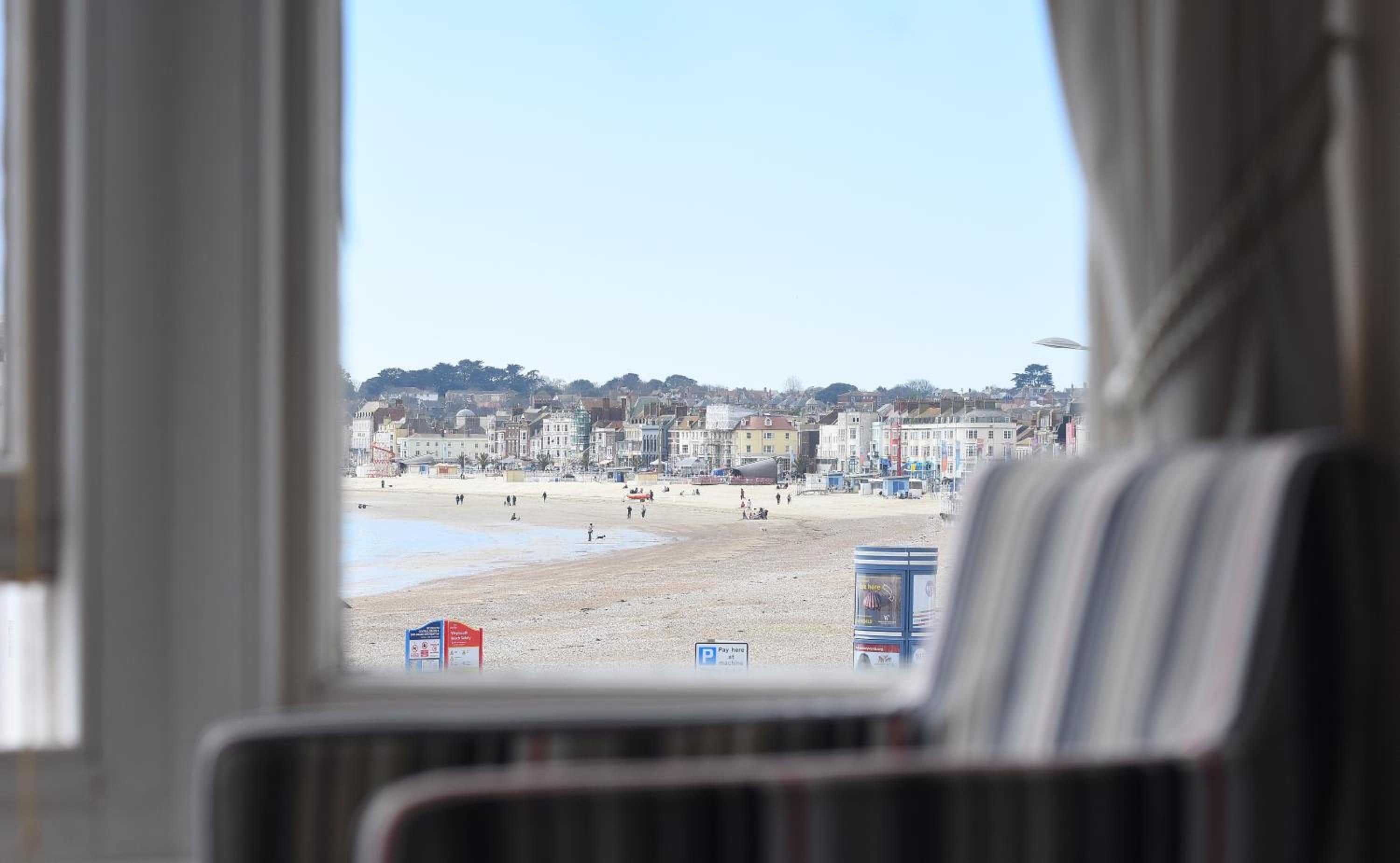 Vista de una playa con edificios al fondo, visible a través de una ventana con cortinas.