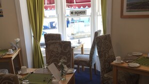 Un coin repas avec une table dressée pour deux personnes, des chaises, une fenêtre sur laquelle est affiché un panneau indiquant « COMPLET » et un présentoir à menus.