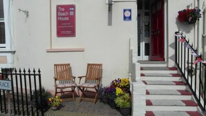 A building with a sign for 'The Beach House' indicating en-suite rooms and sea views. There are two wooden chairs outside, a red door, and a staircase with red and white steps. - The Beach House (Weymouth)