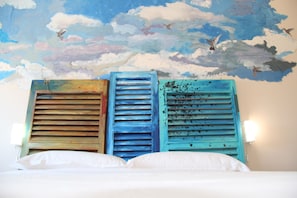 A bed with white linens, a mural of clouds and birds on the wall, and three blue shutters. - Urban Pop Hostel & BnB (Catania)
