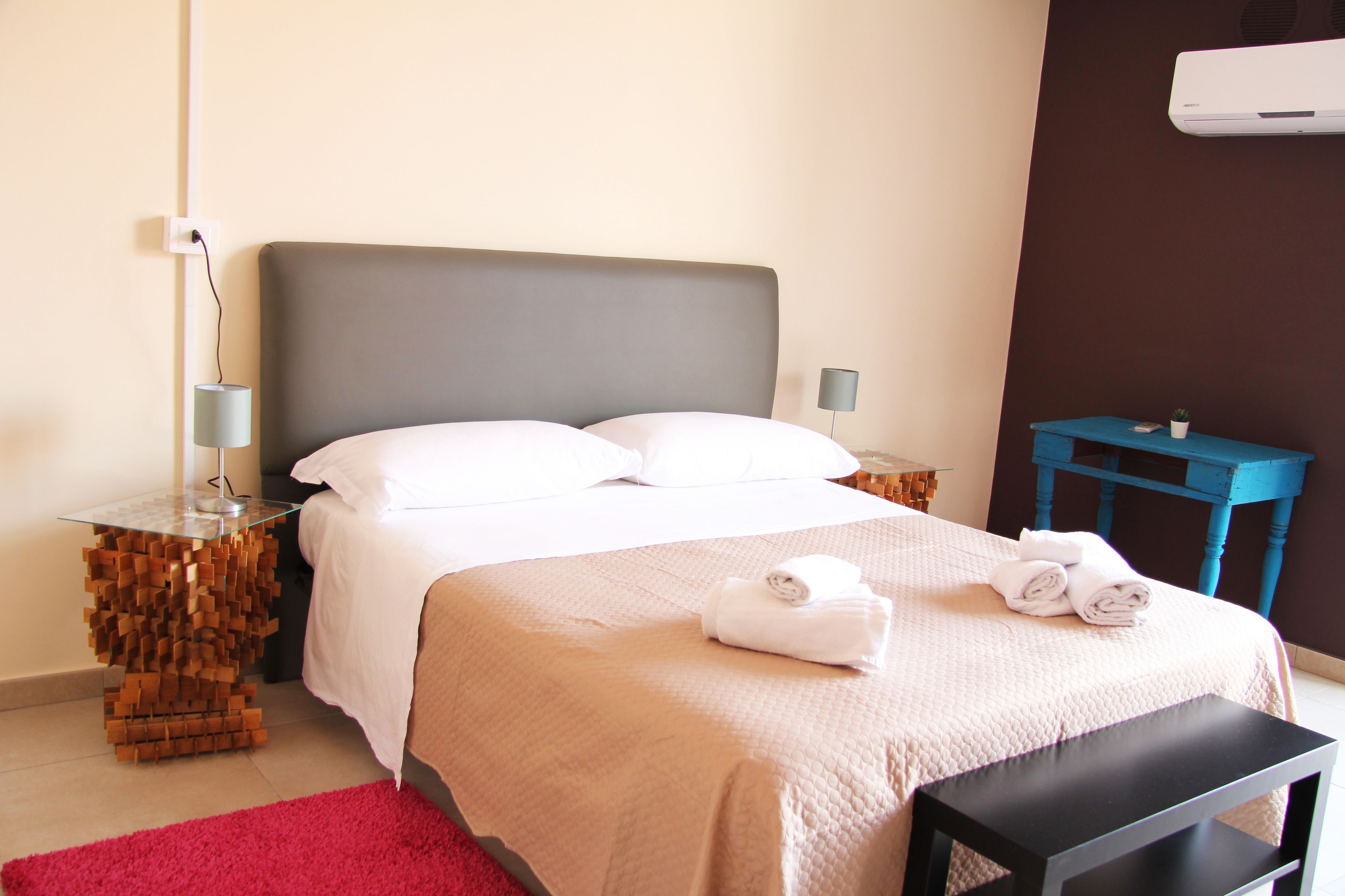 A neatly made bed with white linens, a red rug, and a small table with a plant.
