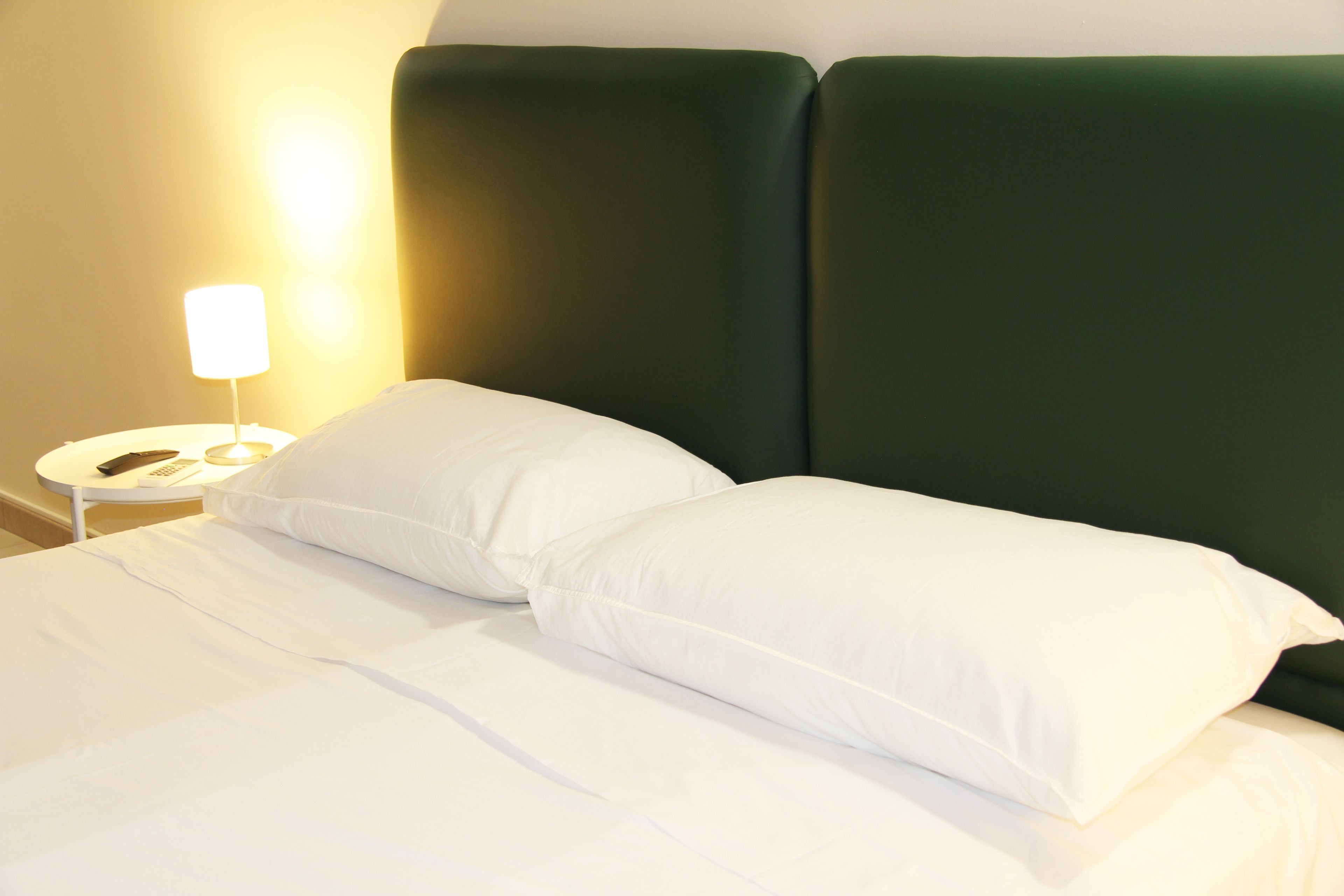 A bed with white linens and pillows, a bedside table with a lamp, and a green headboard.