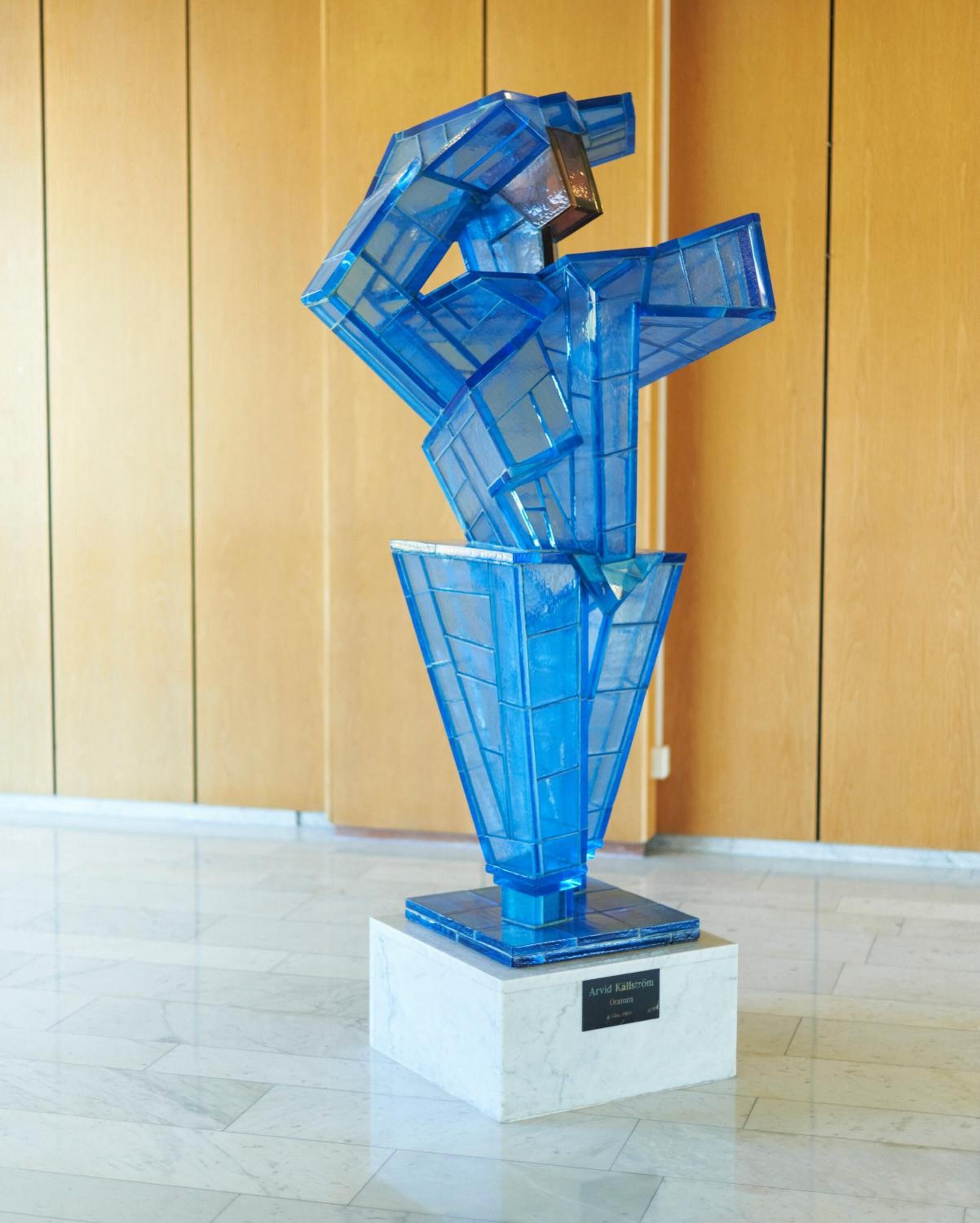 A blue glass sculpture on a marble pedestal in a lobby with wooden paneling.