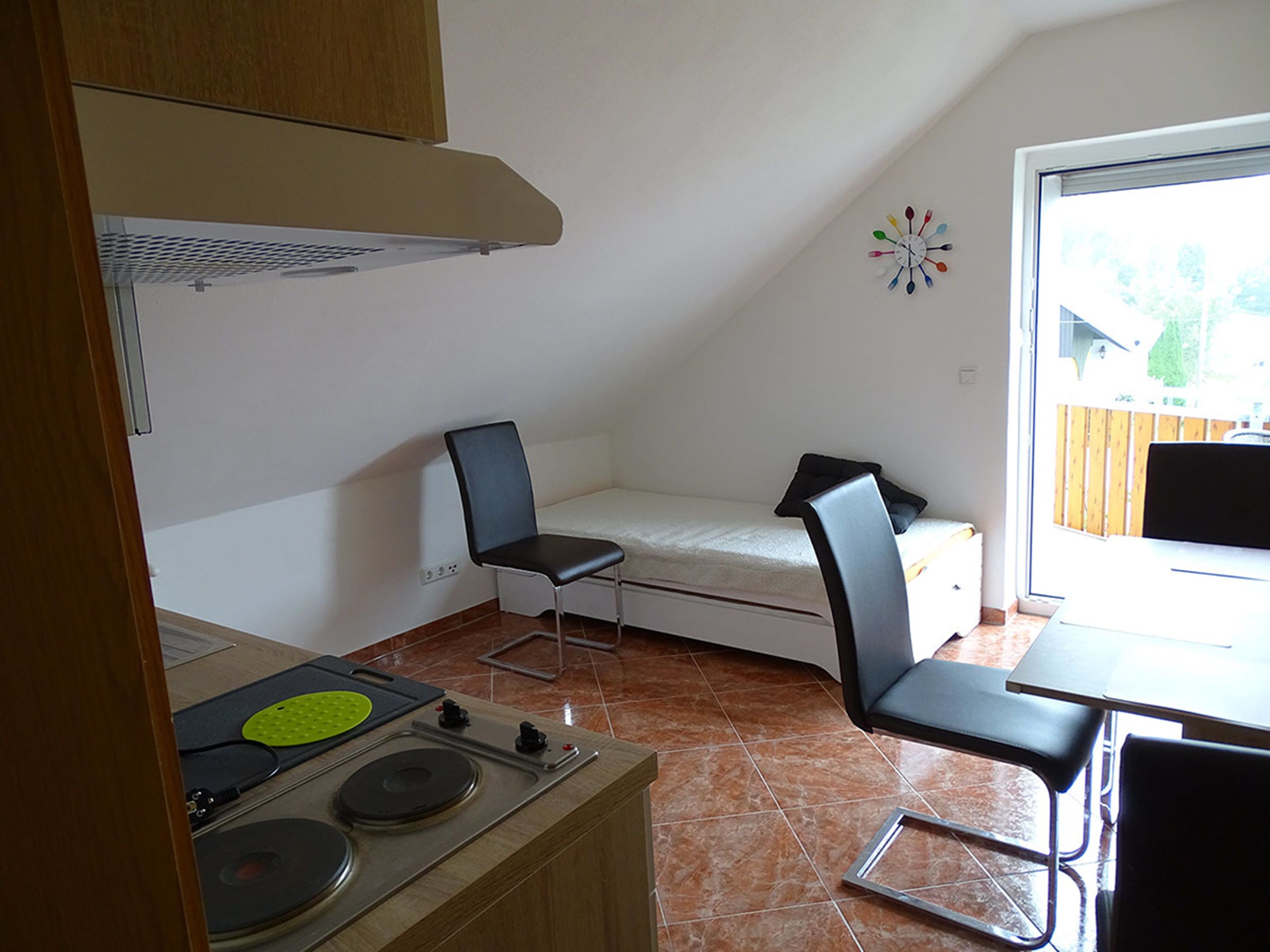 A compact kitchen with a stove, a small dining table, and a chair. There is a bed with a pillow and a wall clock.