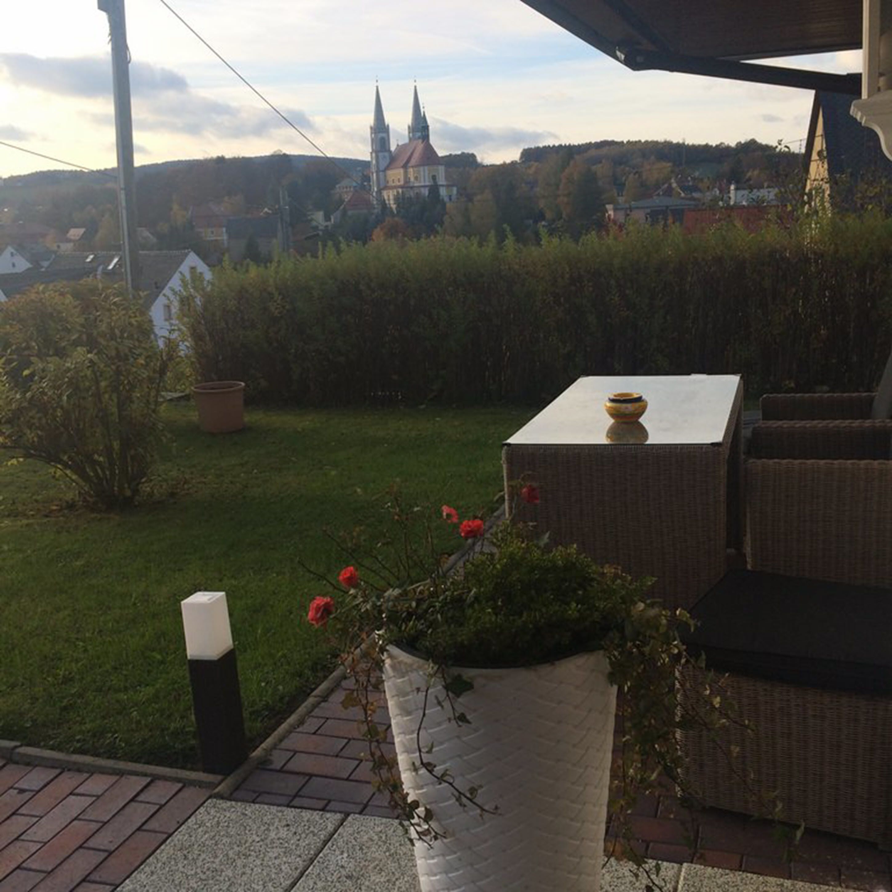 Un patio con mobiliario de mimbre, una maceta con flores rojas y una vista de una iglesia y casas a lo lejos.