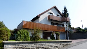 A modern two-story house with a stone retaining wall, a wooden balcony, and a small garden area. - Gästezimmer & Ferienwohnung Berge (Schirgiswalde-Kirschau)