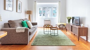 Un salon comprenant un canapé, deux tables d’appoint, un tapis vert, un téléviseur et une fenêtre donnant sur des immeubles.