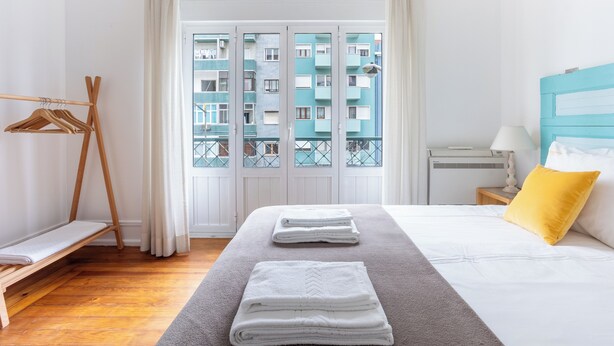 Une chambre d’hôtel avec un lit, un porte-manteau en bois, une fenêtre donnant sur un immeuble, et une table de chevet avec une lampe et un oreiller.