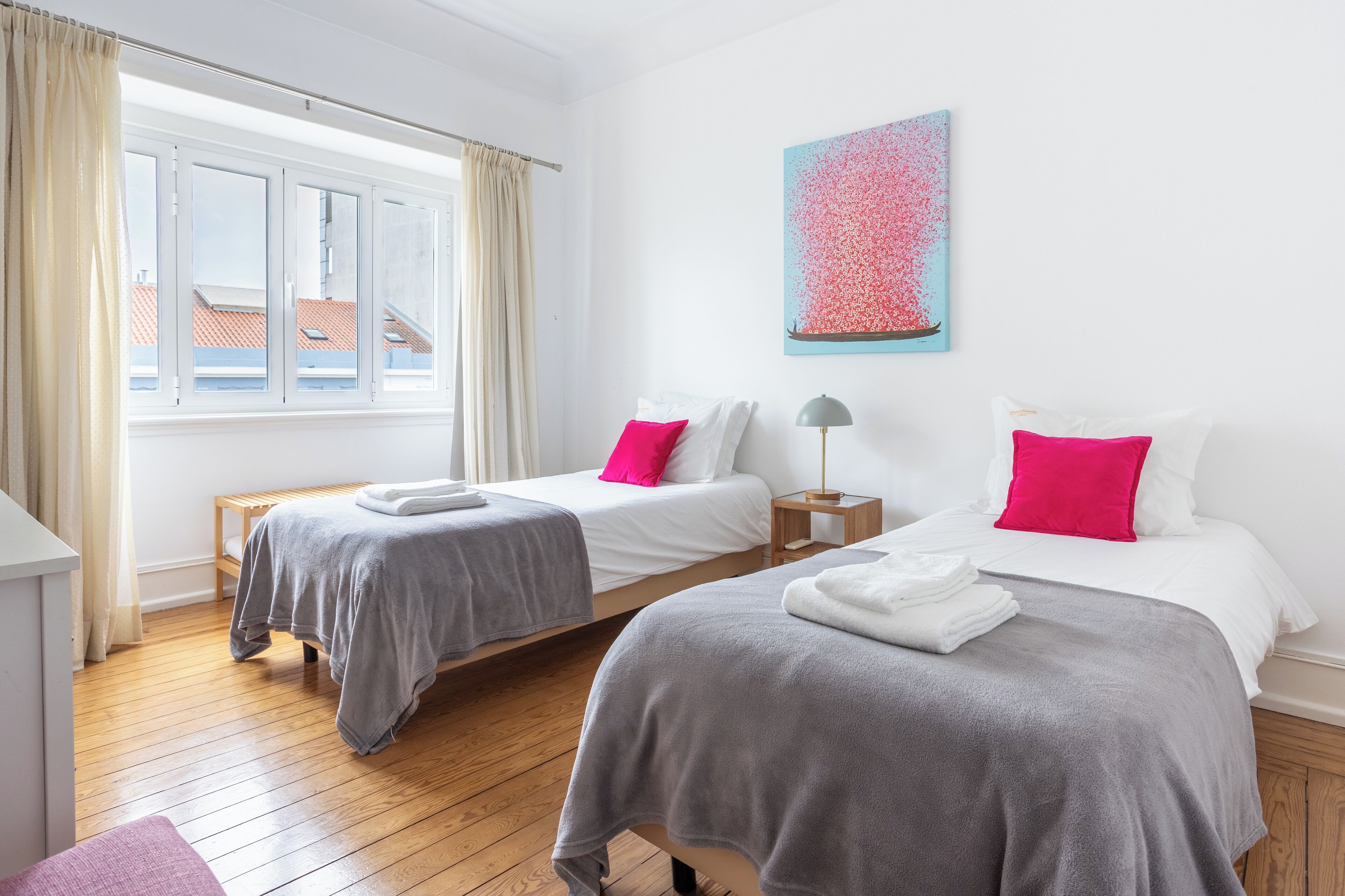 A hotel room with two beds, a window with curtains, a wooden floor, and a painting on the wall.