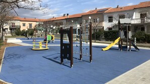 A playground with various play structures, including a slide and climbing frames, surrounded by a paved area and residential buildings in the background. - Apartments Michael (Omisalj)