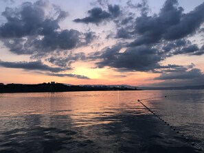 Vista al frente del agua con un muelle que se adentra en el agua, una costa distante con edificios y un cielo de atardecer con nubes.