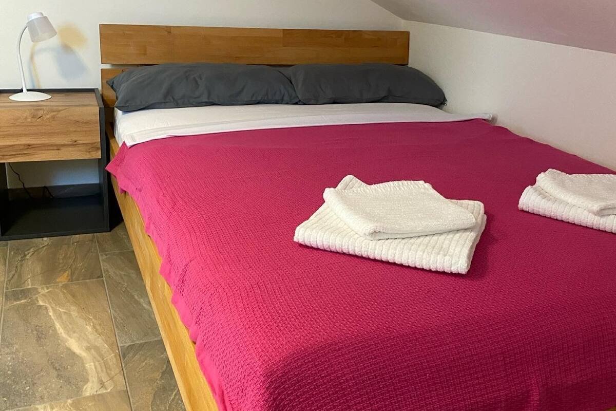 A single bed with a wooden headboard, a pink blanket, two white towels, and a gray pillow.