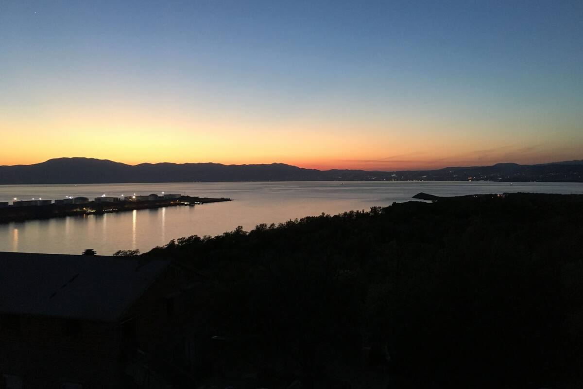 A coastal landscape during sunset with a calm sea, distant mountains, and a small settlement with buildings and lights.