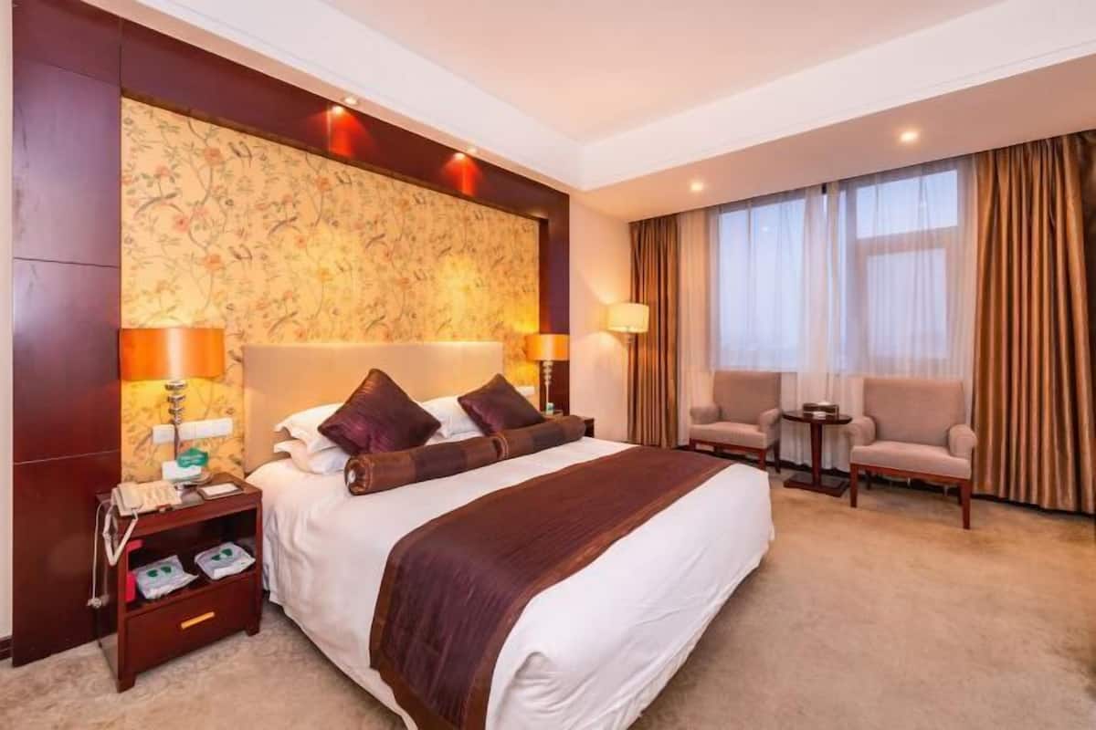 Quarto de hotel com uma cama grande, duas luminárias de cabeceira, uma mesinha de cabeceira, uma pequena mesa, duas poltronas e uma janela grande com cortinas.
