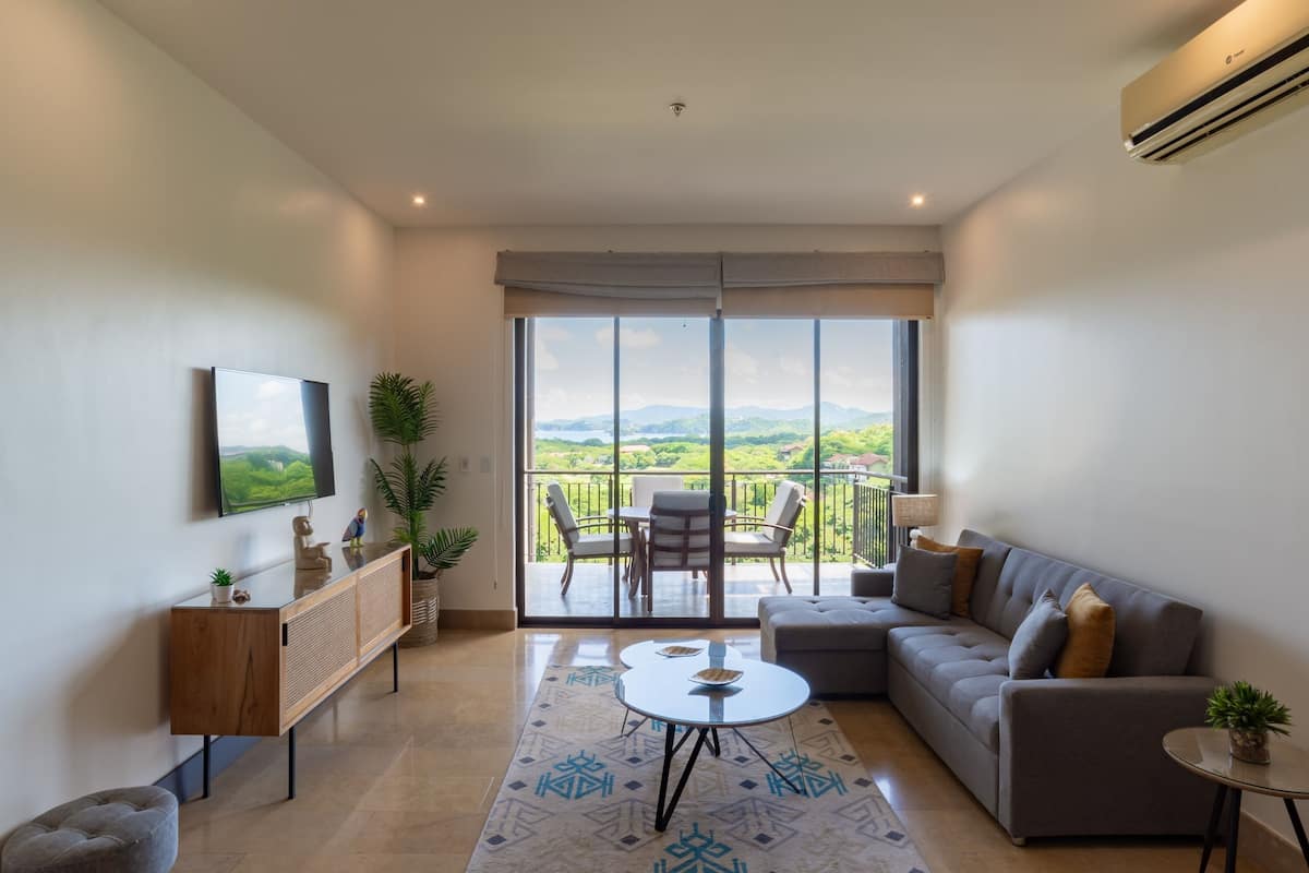 A modern living room with a sofa, a coffee table, a TV mounted on the wall, and a balcony with a view of the countryside.