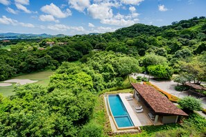 A poolside area with a long rectangular pool, a covered seating area with lounge chairs, and a view of a lush green landscape with trees and hills in the background. - Reserva Conchal Resort - Roble Sabana Complex (Cabo Velas)