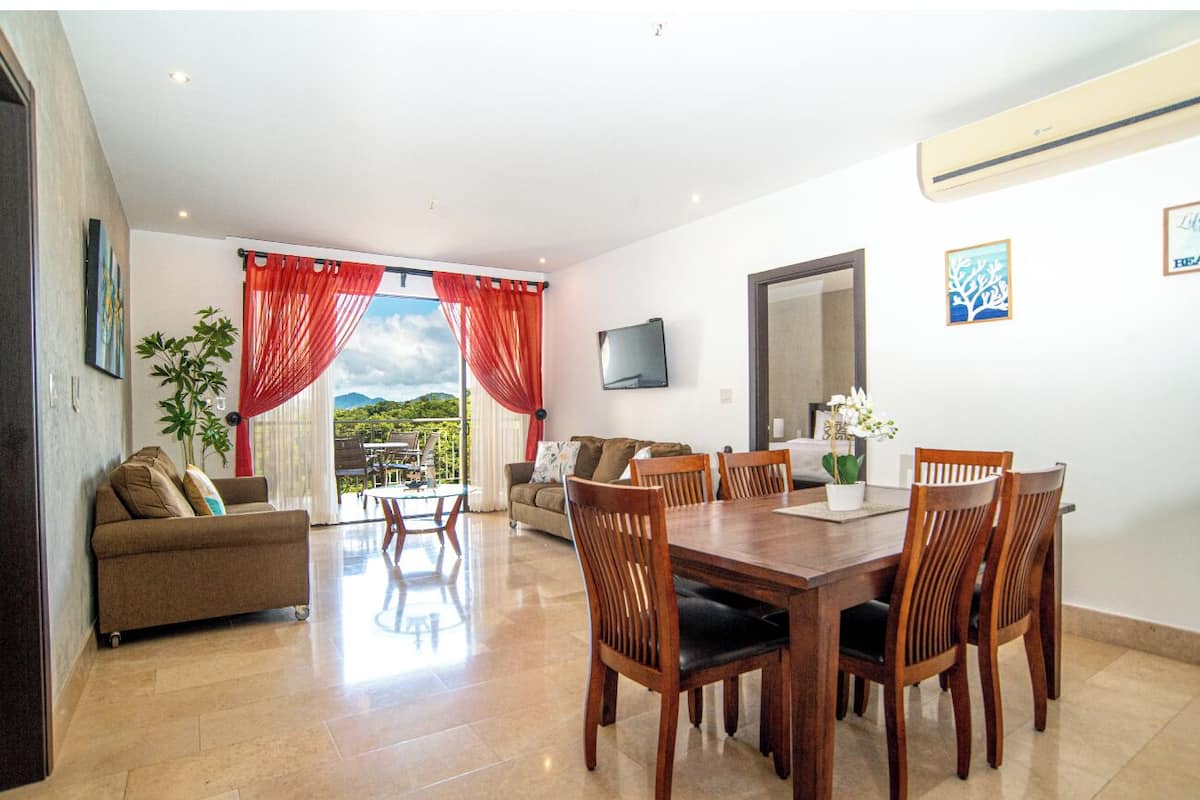 A spacious living area with a dining table and chairs, a sofa, and a TV mounted on the wall.