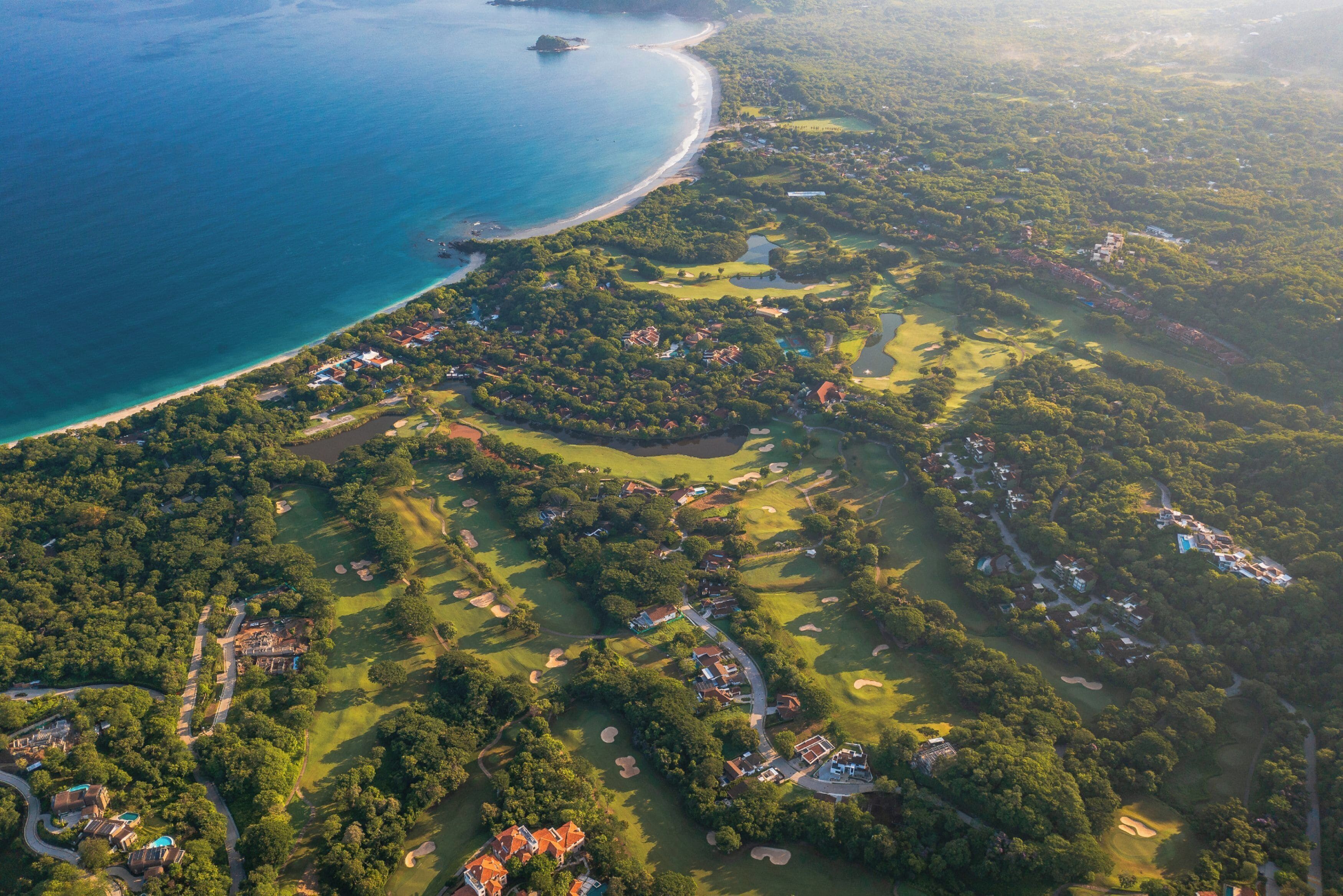 Luftaufnahme einer Küstenregion mit einem Golfplatz, Strandimmobilien und dichter Vegetation.