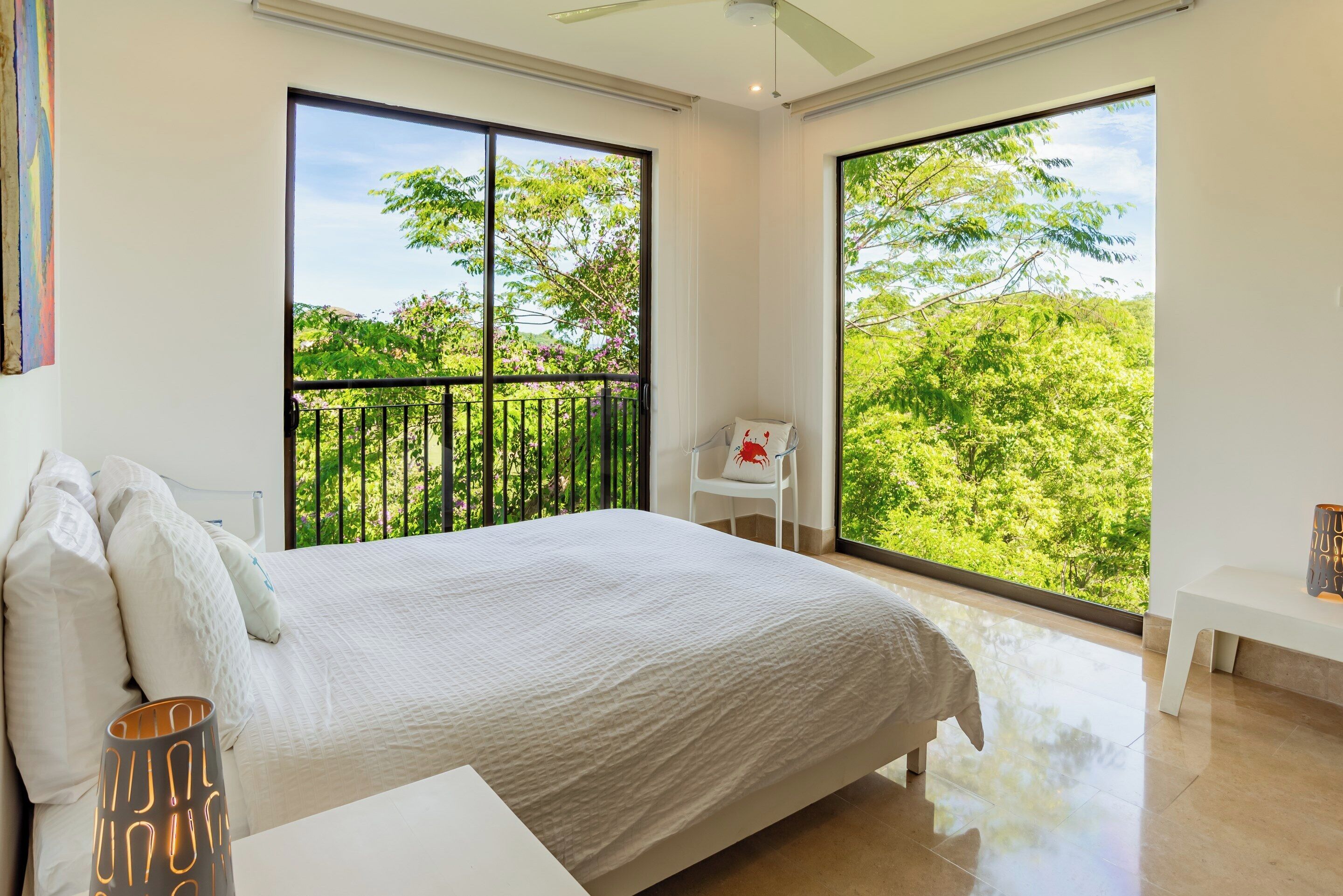 A bedroom with a large bed, a white chair, and a view of greenery through large windows.