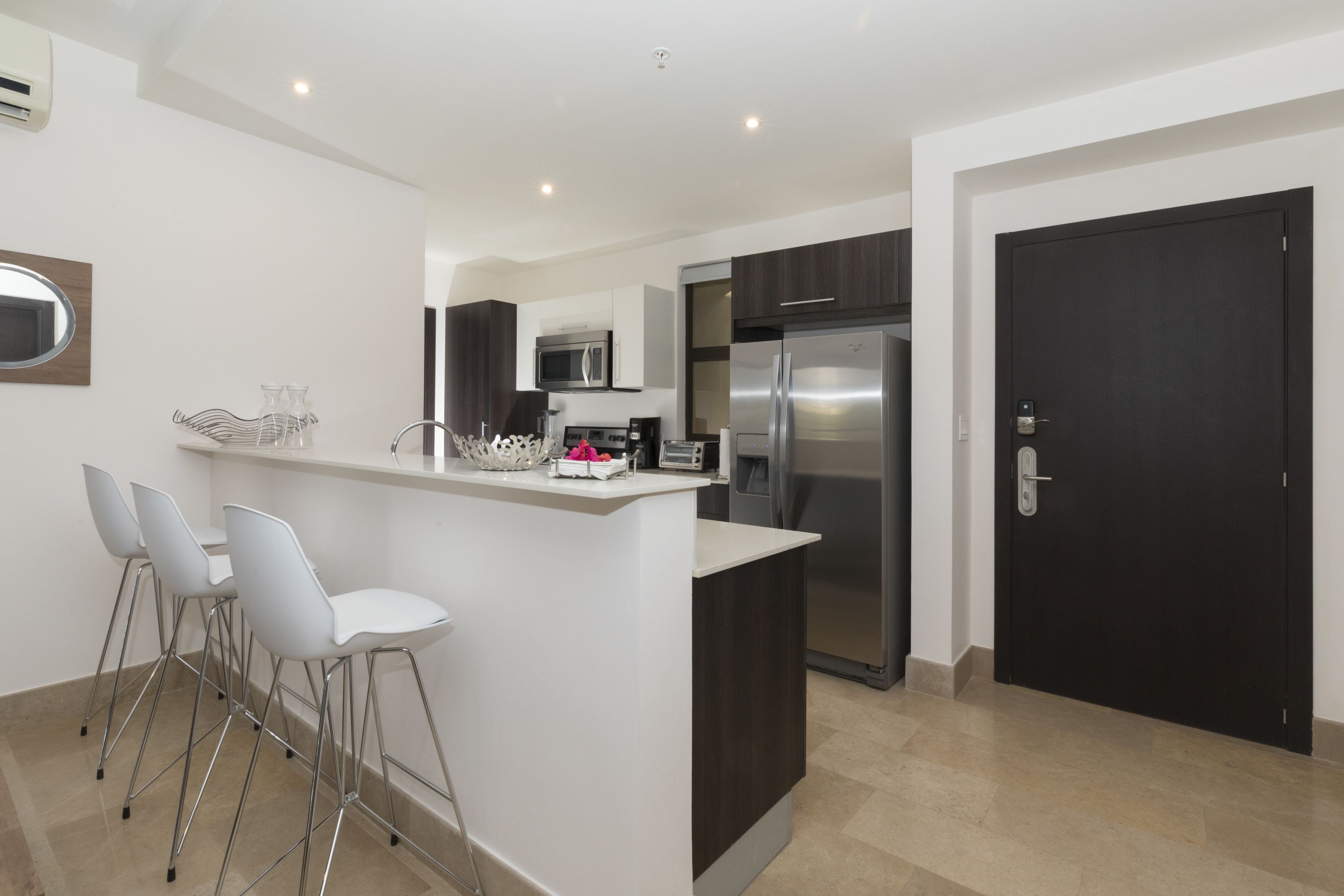 A modern kitchen with a central island, bar stools, a microwave, a refrigerator, and a dark door.