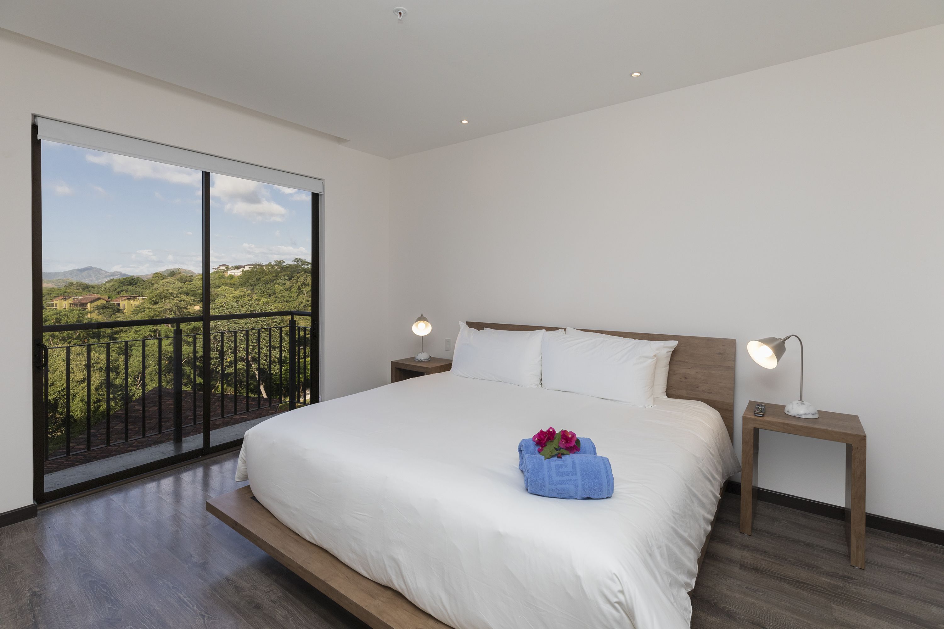 A bedroom with a large bed, two bedside tables with lamps, a view of greenery through a balcony door, and a wooden floor.