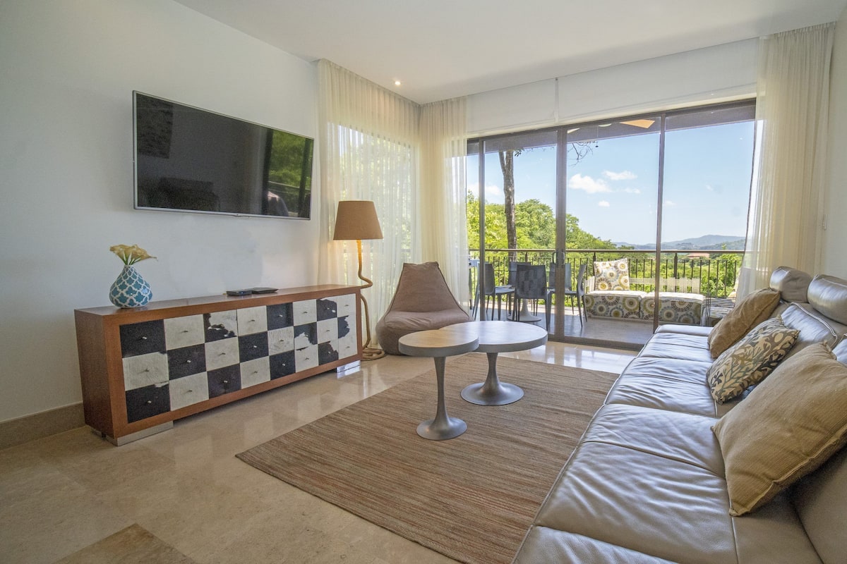 A modern living room with a sofa, a coffee table, a TV, and a balcony with outdoor seating.