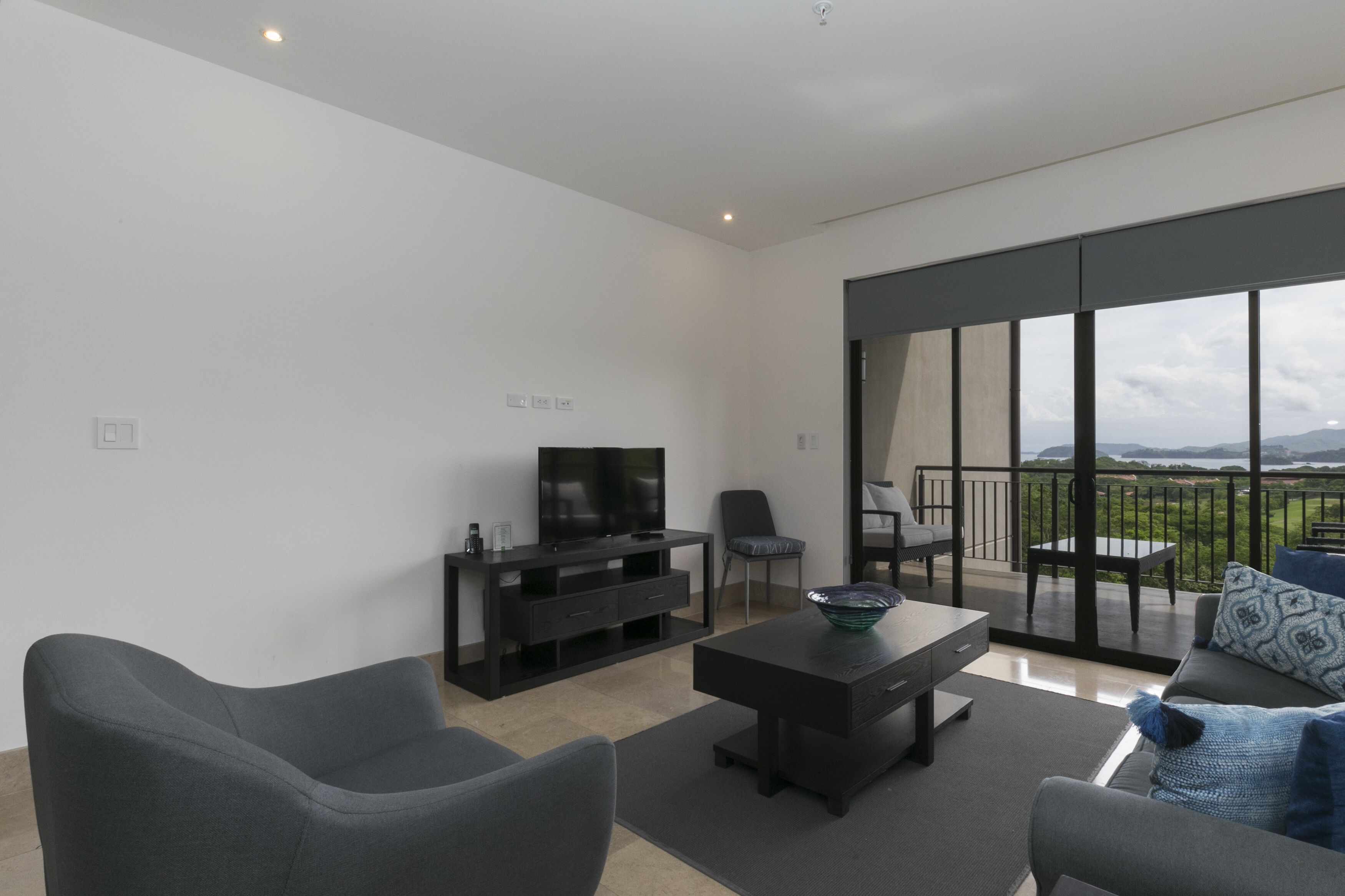 A modern living room with a flat-screen TV, comfortable seating, a coffee table, and a balcony with a scenic view.