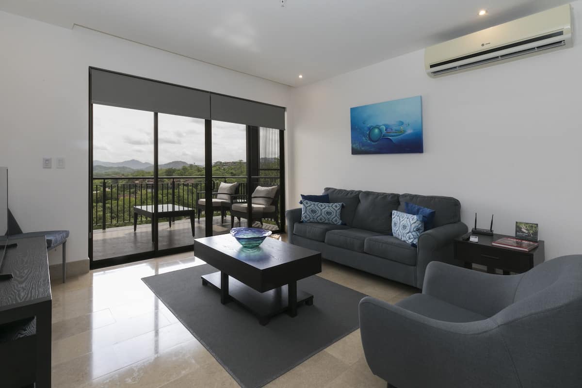 A modern living room with a sofa, coffee table, and chairs, a balcony with a view of the mountains, and an air conditioning unit on the wall.